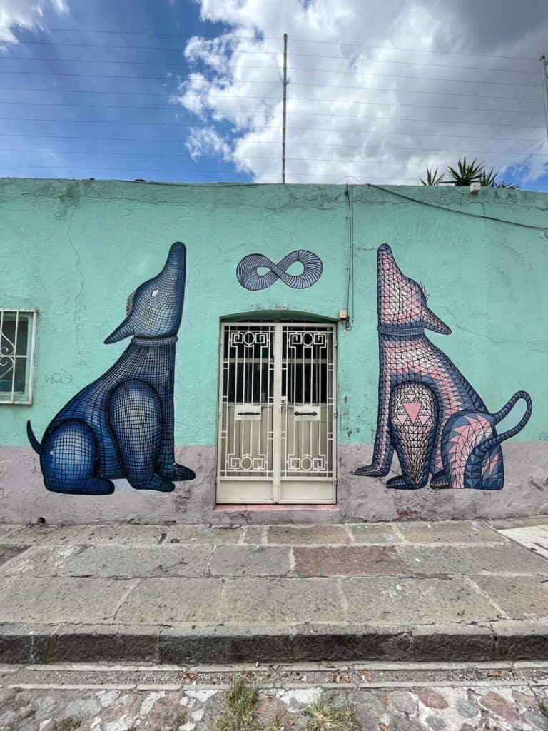 A pale turquoise building with a large blue dog painted on one side of the door and another pale pink dog on the other side both looking up howling in Xanenetla area of Puebla.