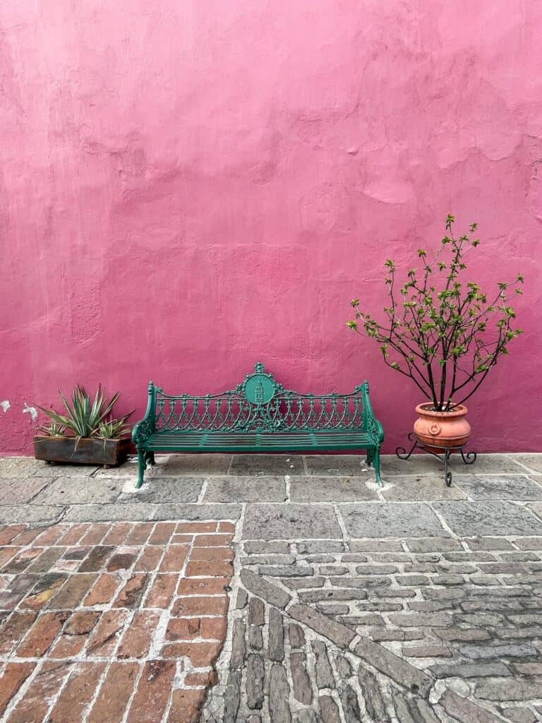 A beautiful vibrant pink wall with a green iron bench and two pots with plants on either side along a brick walkway in Puebla.