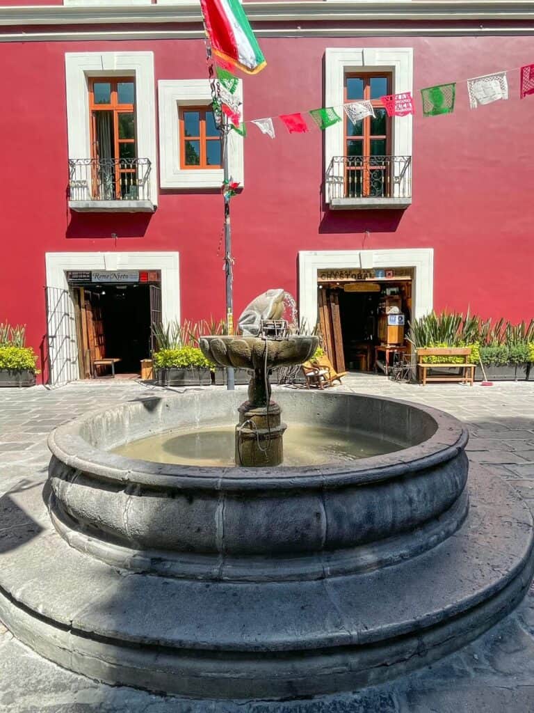 Water fountain with water spouting out of a frog with a dark red building with white trim behind it on Alley of the Frogs.