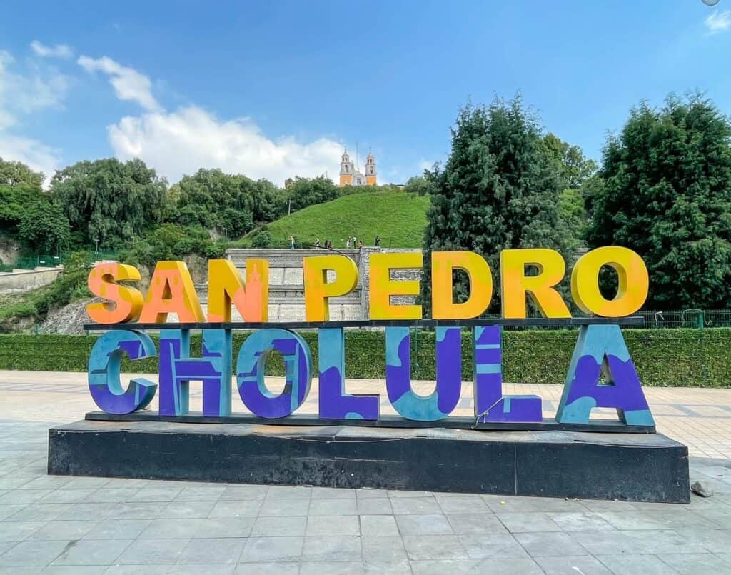 Huge letters in yellow and blue that spell out San Pedro Cholula at the base of the Cholula Pyramid and the yellow church at the top.