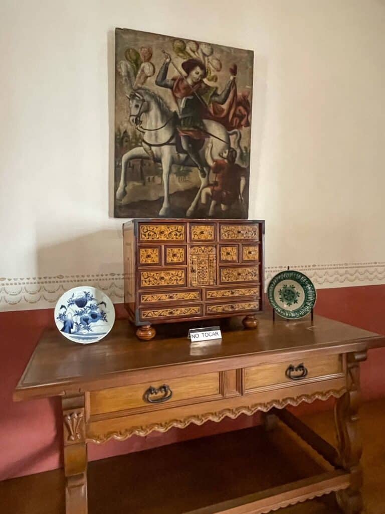 A painting of a man on a horse above and beautiful wooden desk, plates with artwork, and a small chest with drawers.