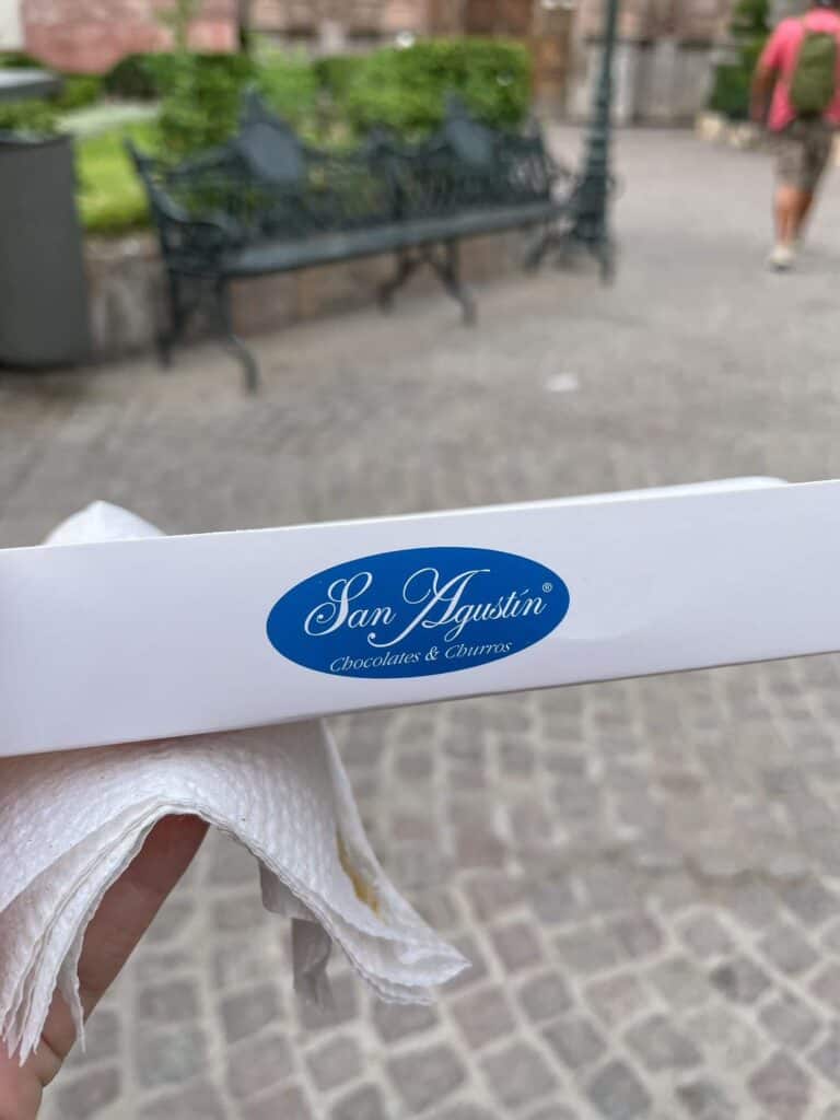 Holding a small, thin paper container that fits a churro perfectly and has the blue and white logo of San Agustin churros on it.