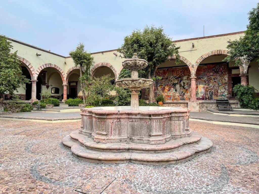 A stone water fountain in the center of the outdoor courtyard at the Instituto Allende with trees, art murals, and arched walkway behind.