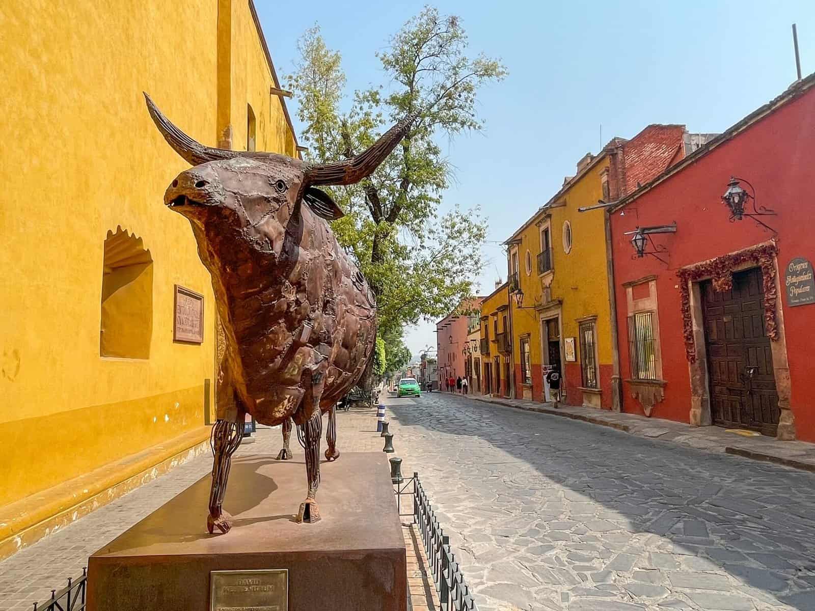 A bronze statue of a steer in front of a bright yellow building and a charming cobblestoned street in San Miguel de Allende, Mexico.