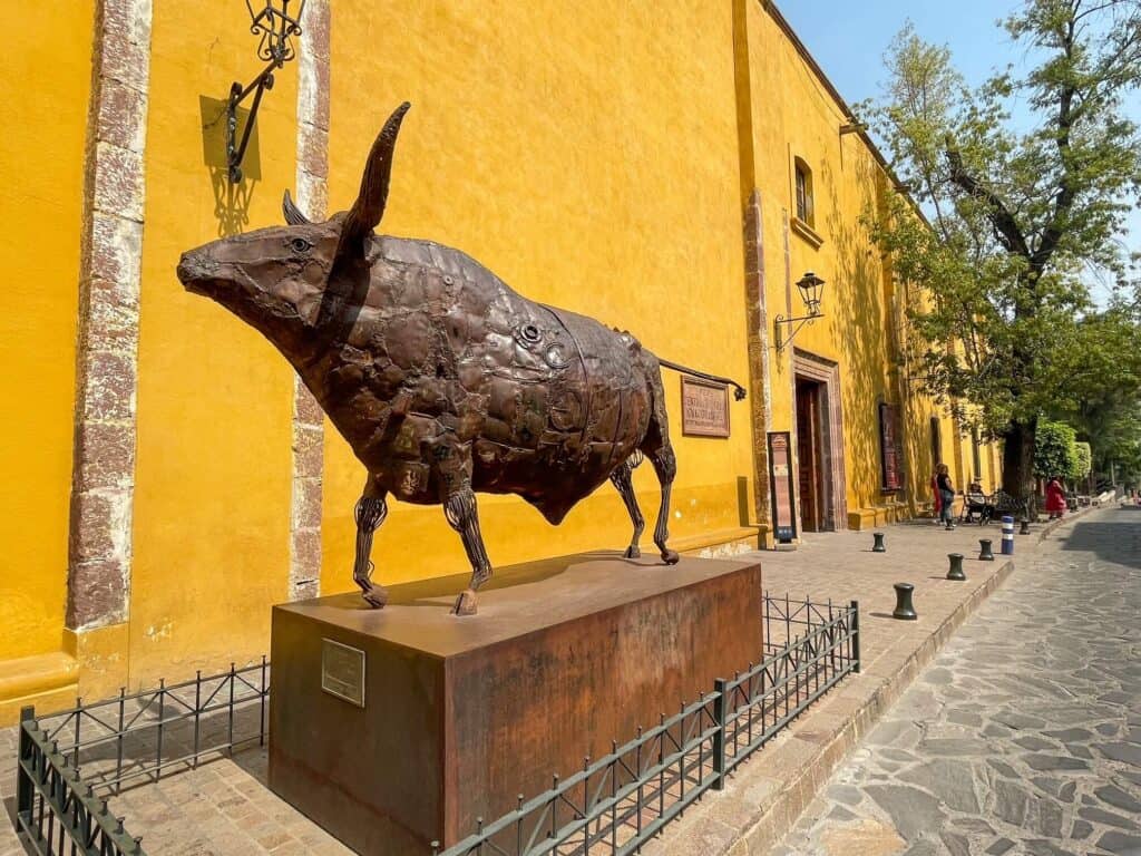 A bronze statue of a bull in front of the bright yellow building of the Centro Cultural Ignacio Ramirez in San Miguel de Allende, Mexico.