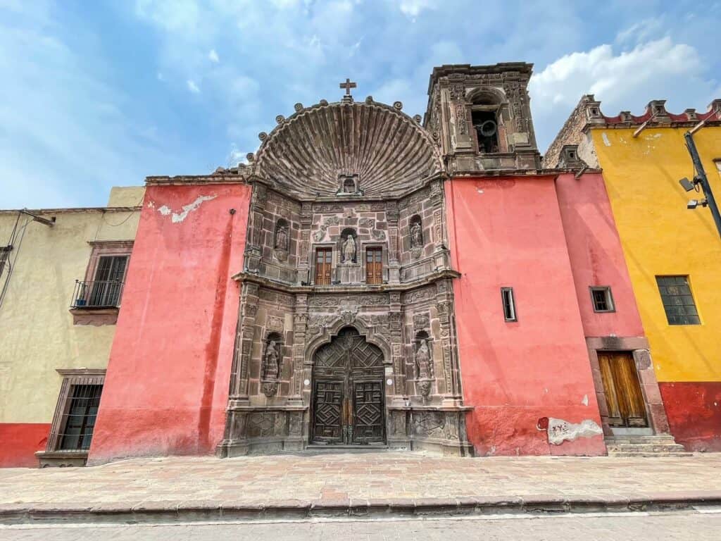 The beautiful stone carving facade of the entrance to one of many beautiful churches with pink walls on the sides and a tower on the right side in Plaza Civica in San Miguel de Allende.