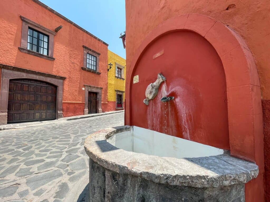 Walking around a corner of a street with red and yellow colored building is a small water spout with a red background and stone basin where you can fill up a bottle with water in San Miguel de Allende.