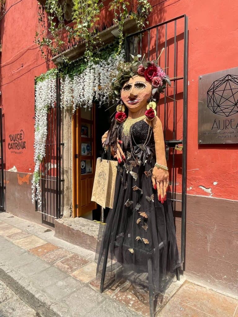 White delicate flowers hanging over over a doorway with a large paper mache woman in traditional Mexican dress in front of a shop in the historic San Miguel de Allende.
