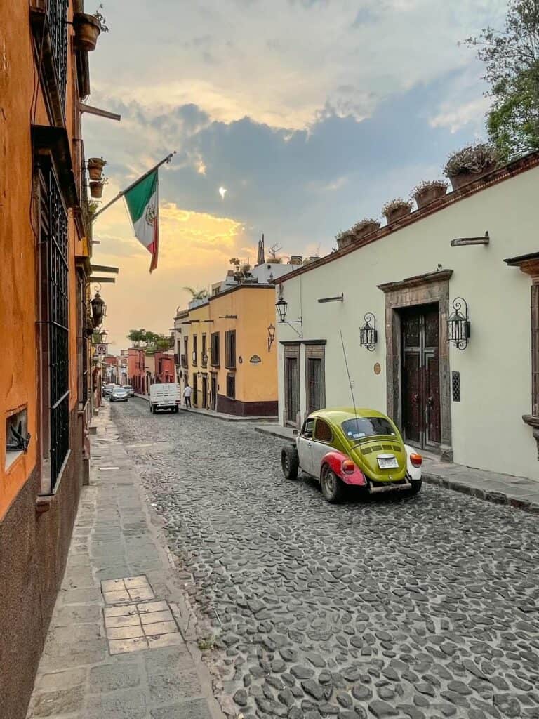 Walking on a cobblestoned street with colorful buildings, a Mexican flag hanging, and a VW bug driving past in colors of red, green, and red close to sunset in San Miguel de Allende.