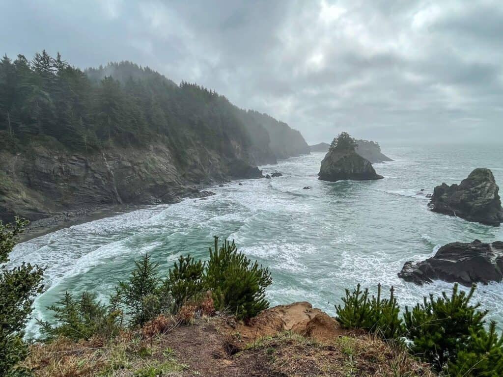 Looking south at the tall cliffs, many large rock formations off the shore, emerald water and fog at Spruce Island Viewpoint in Southern Oregon.