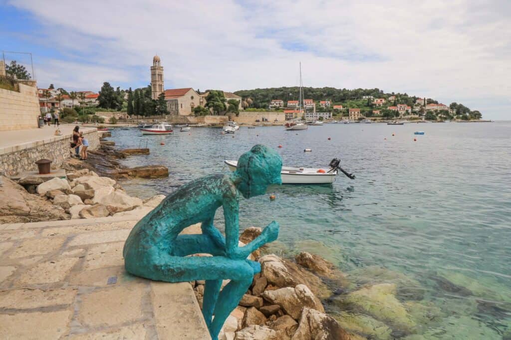 A teal green statue of a woman leaning over with her arms on her thighs at the edge of the marina with clear blue water and a church tower in the distance on Hvar Island near Split, Croatia.