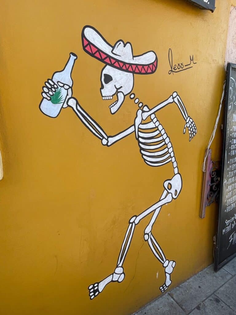 An art mural of a skeleton wearing a sombrero hat and holding a bottle of mezcal with a yellow background in Oaxaca City, Mexico.