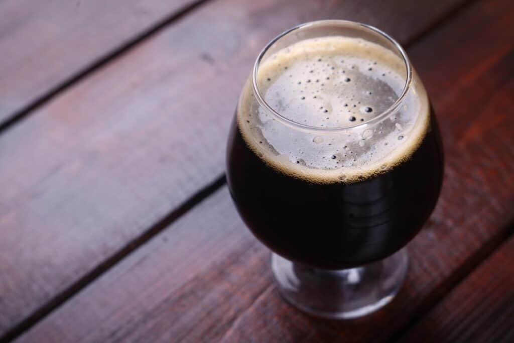 A snifter glass full of dark stout ale on a dark wooden table in a brewery.