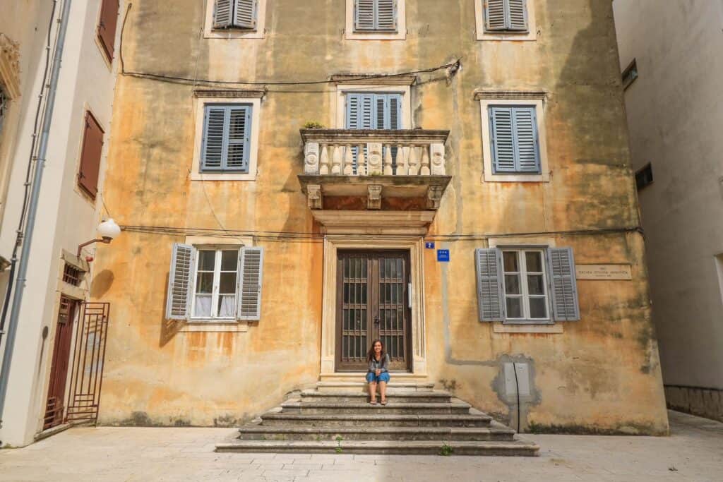 Sitting on the front steps of an old but pretty building in Zadar that is in shades of yellow and green.