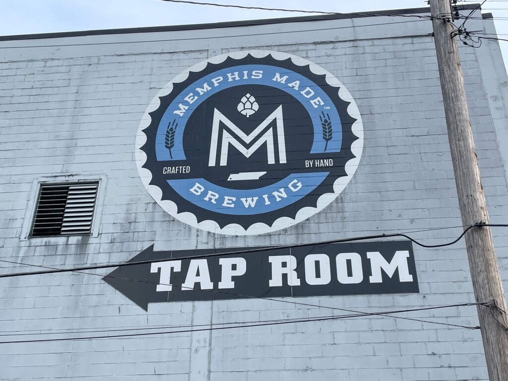 The Memphis Made Brewing logo on the side of the building in shades of light and dark blue with an arrow pointing to the tap room entrance in Memphis.