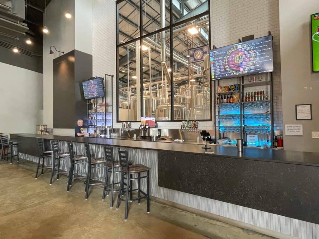 The bar with tall chairs at Crosstown Brewing Co with a large window looking into the brewery and large stainless steel tanks.