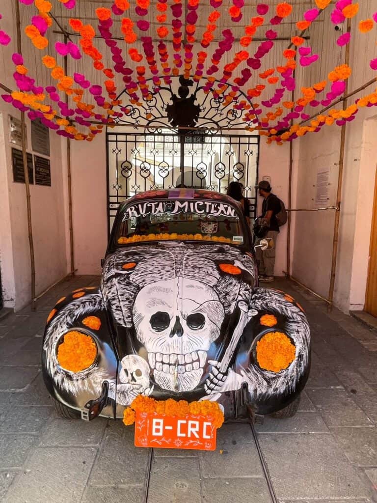 A VW bug car painted in a Day of the Dead theme with a huge skeleton face on the front and flowers hanging from above.