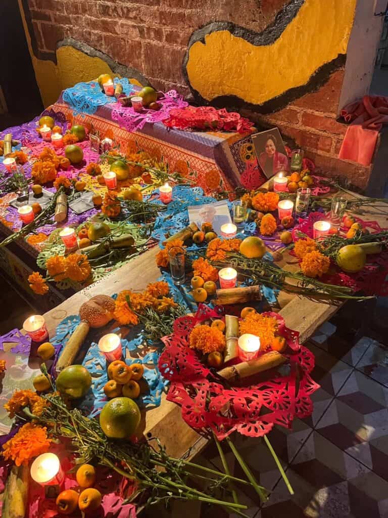 A close up of an altar with several different photos, candles, fruit, flowers, and mezcal honoring those who have passed during a Day of the Dead tour experience.