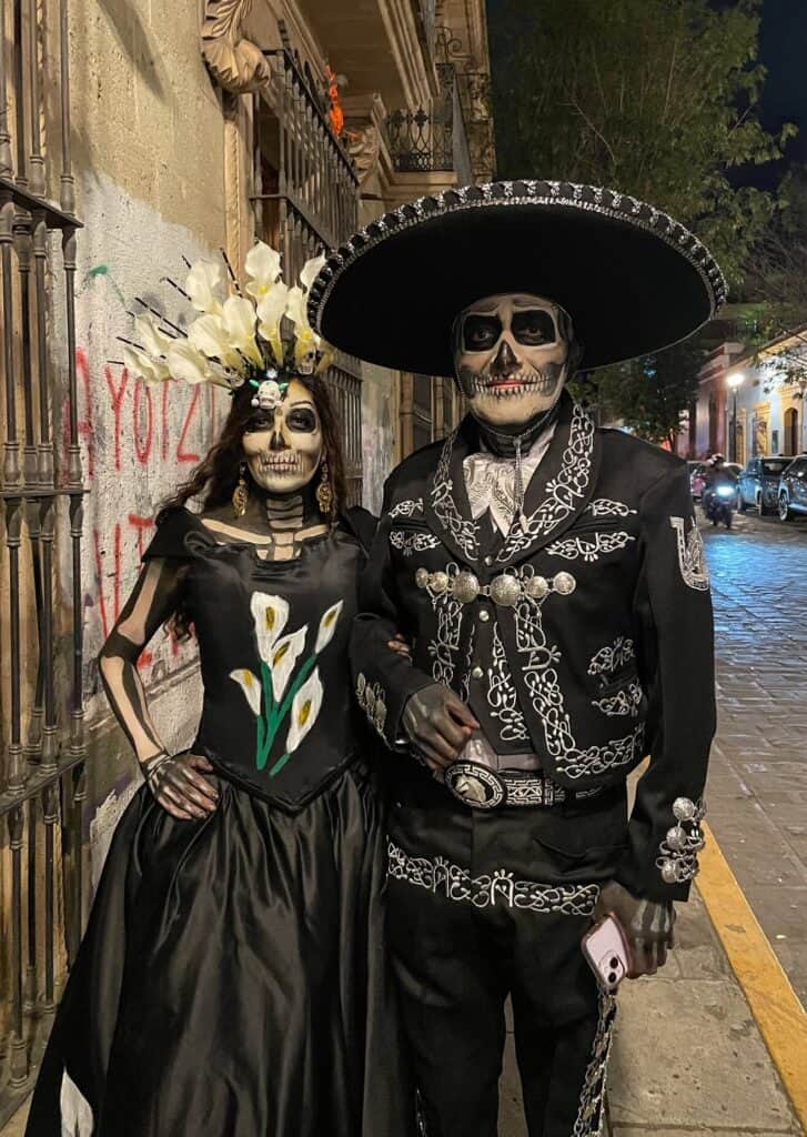 A local couple dressed all in black traditional attire with incredible skeleton face paint and on their arms and hands resembling real skeletons plus a flower headband and a hat on the man in Oaxaca during Dia de los Muertos.