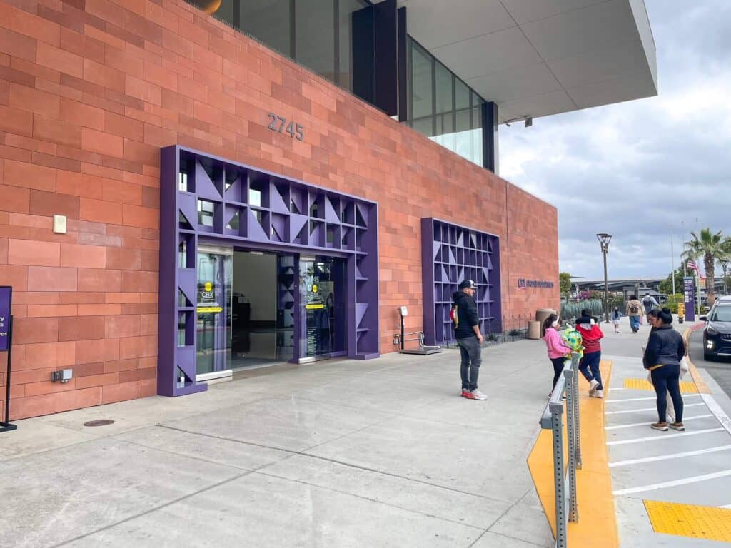 The front entrance to CBX on the American side with its rust colored exterior and deep purple accents and people unloading from the curb.