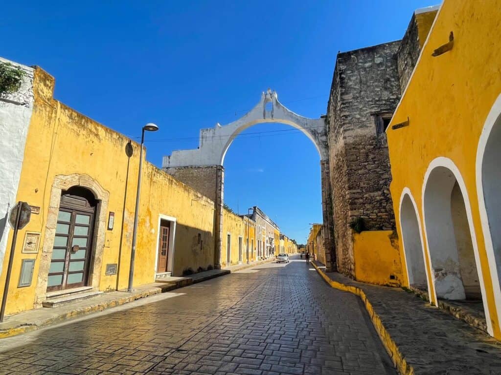 Yellow colored buildings along a cobblestone street and a white arch connecting the two sides of the street in Izamal.
