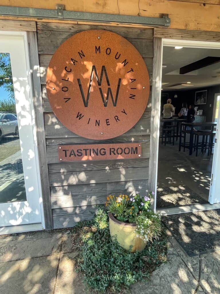 The front entrance into the Volcan Mountain Tasting Room with a sign with the logo and a plant by the door in the hills of Julian.