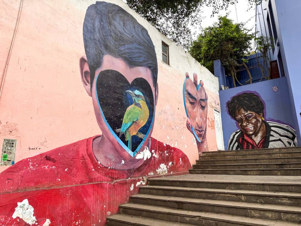 An art mural that is of an abstract art piece in the gallery next door of a boy whose face is in the shape of a heart with a bird painted in the heart and holding up in his hand a heart with his face painted on it on the steps leading up to the Bridge of Sighs.