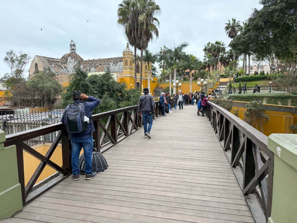 Standing on the Bridge of Sighs in the Barranco neighborhood of Lima with a yellow church and palm trees on the other side.