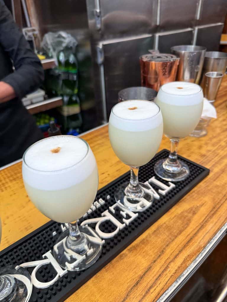 A close up of 3 white colored Pisco Sour cocktails lined in a row on a bar top in Lima.