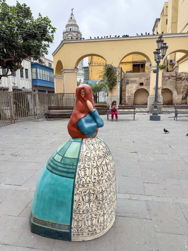 A Las Tapadas Limenas (veiled woman of Lima) statue with a blue and white dress and a brown veil over her head and face with only one eye showing.