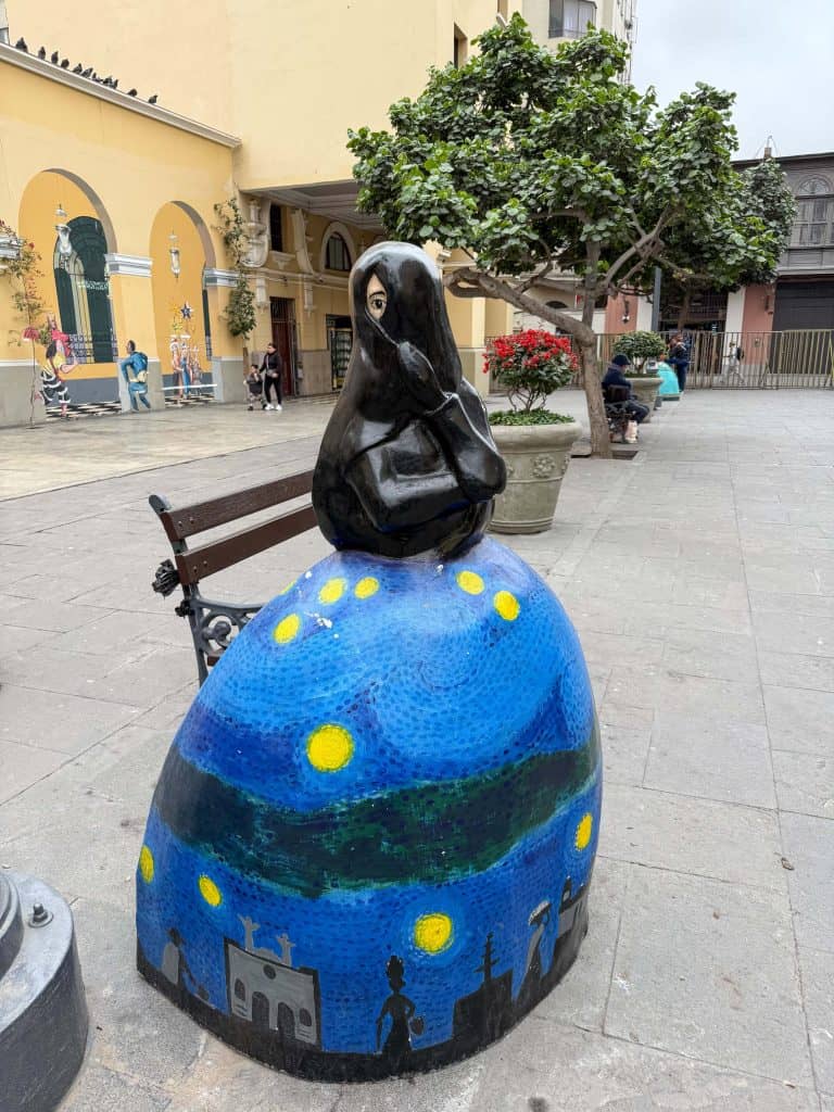A Las Tapadas Limenas (veiled woman of Lima) statue with a dark blue and black dress with yellow stars and a black veil over her head and face with only one eye showing.