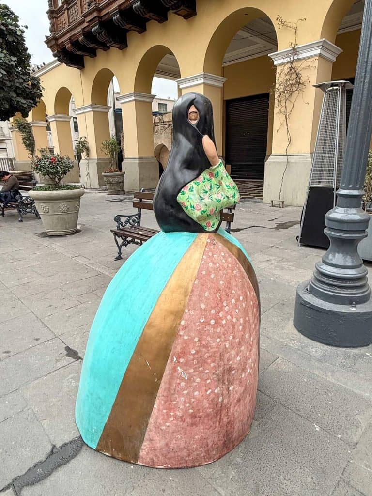 A Las Tapadas Limenas (veiled woman of Lima) statue with a turquoise, gold, and pale pink dress and a black veil over her head and face with only one eye showing.