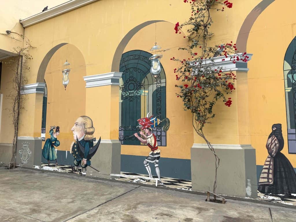 An art mural with various characters against a yellow backdrop and vines with flowers in the historic district of Lima.