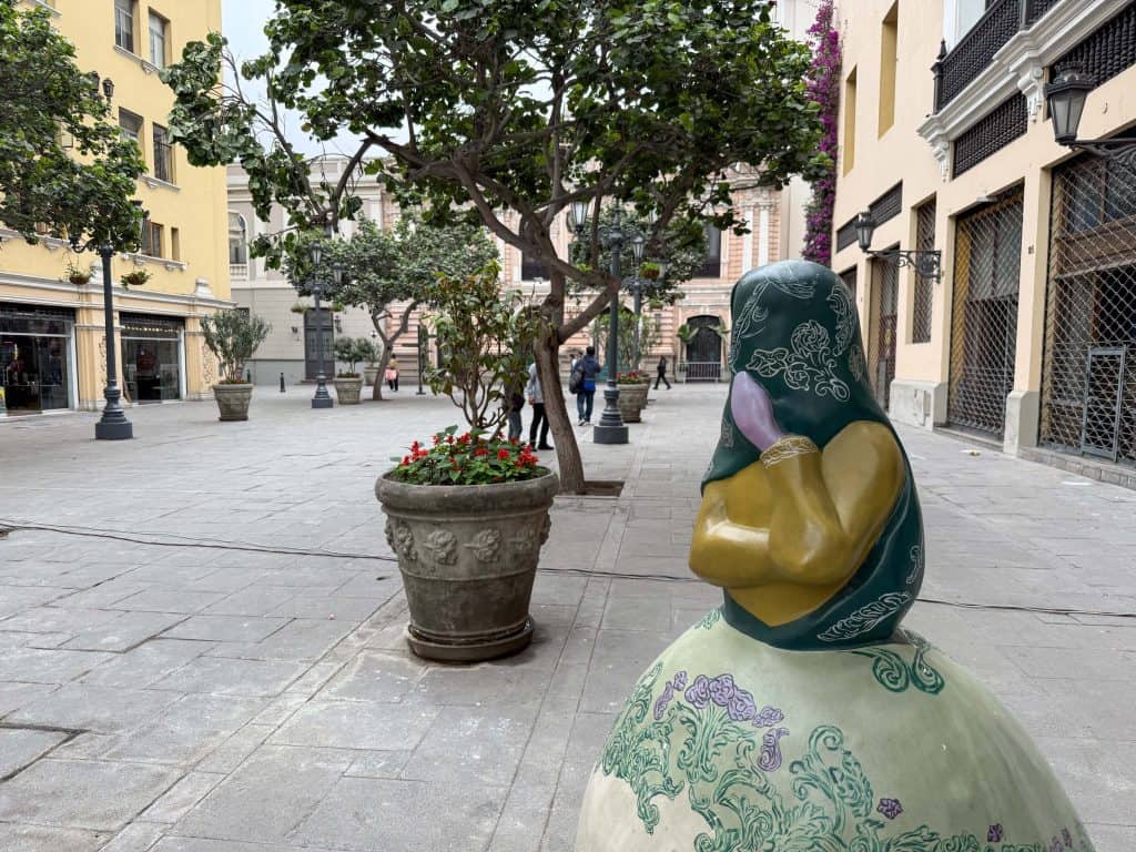 A Las Tapadas Limenas (veiled woman of Lima) statue with a creamy white, gold, and emerald green dress and a deep emerald green veil over her head and face with only one eye showing.