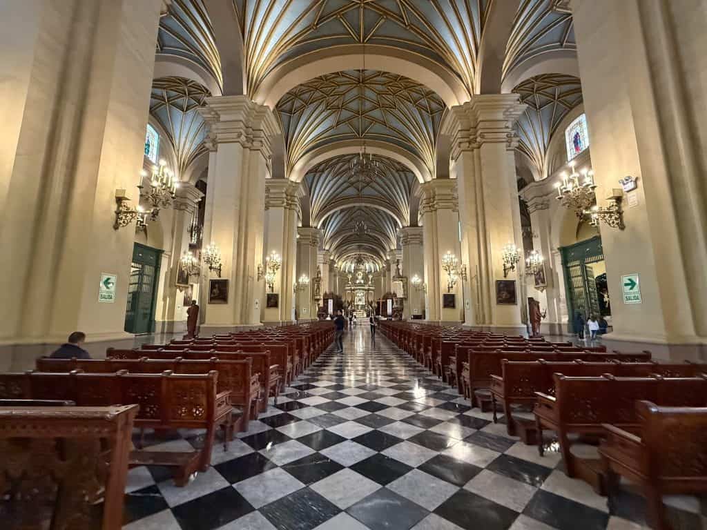 Inside the Lima Cathedral with its ornate architecture, brightly lit, wooden pews, and black and white checkered floor.