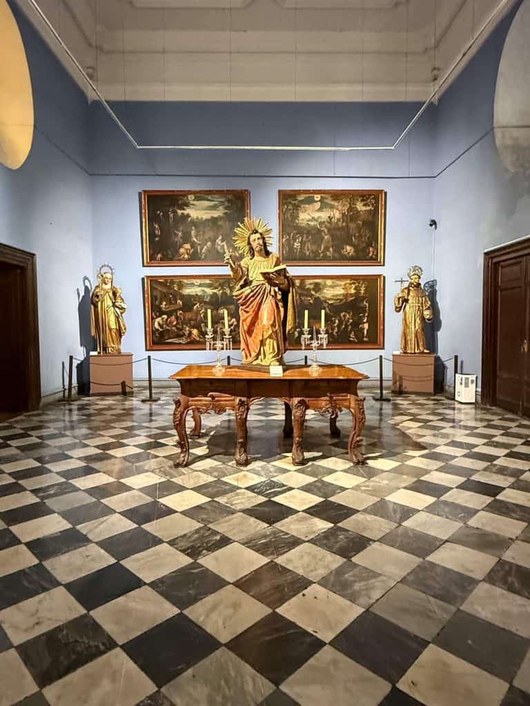 A small room in the Lima Cathedral museum lined with paintings on the pale blue walls, a statue of Jesus in the center and black and white checkered flooring.