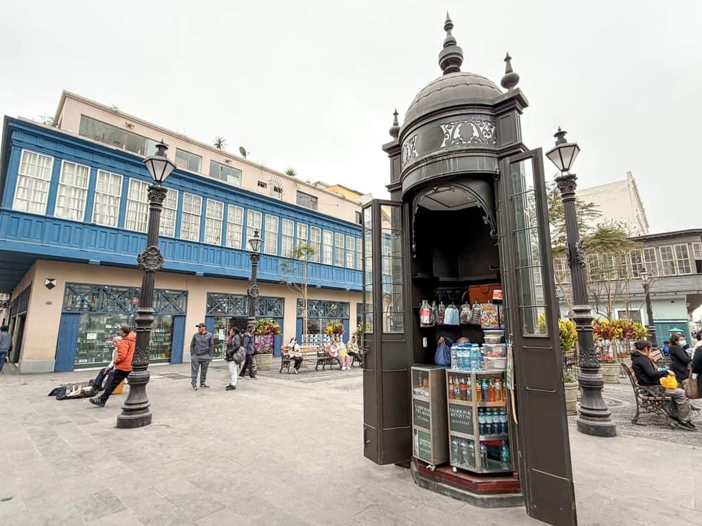 A small square near Plaza de Armas in the historic district with a building with blue box balconies, benches, a small vendor, and old lamp posts in Lima.