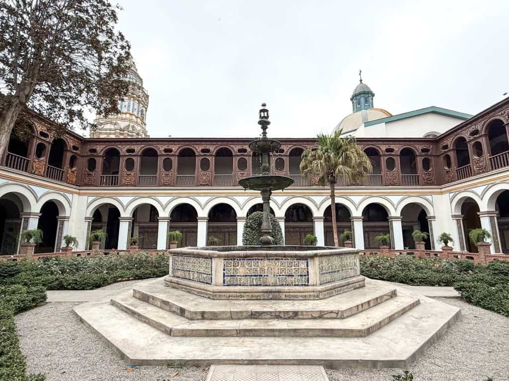 View of the inner courtyard at Santa Domingo convent with a fountain in the center, rose bushes around it and the arched cloisters surrounding the courtyard with the top of the bell tower seen on the far left side.