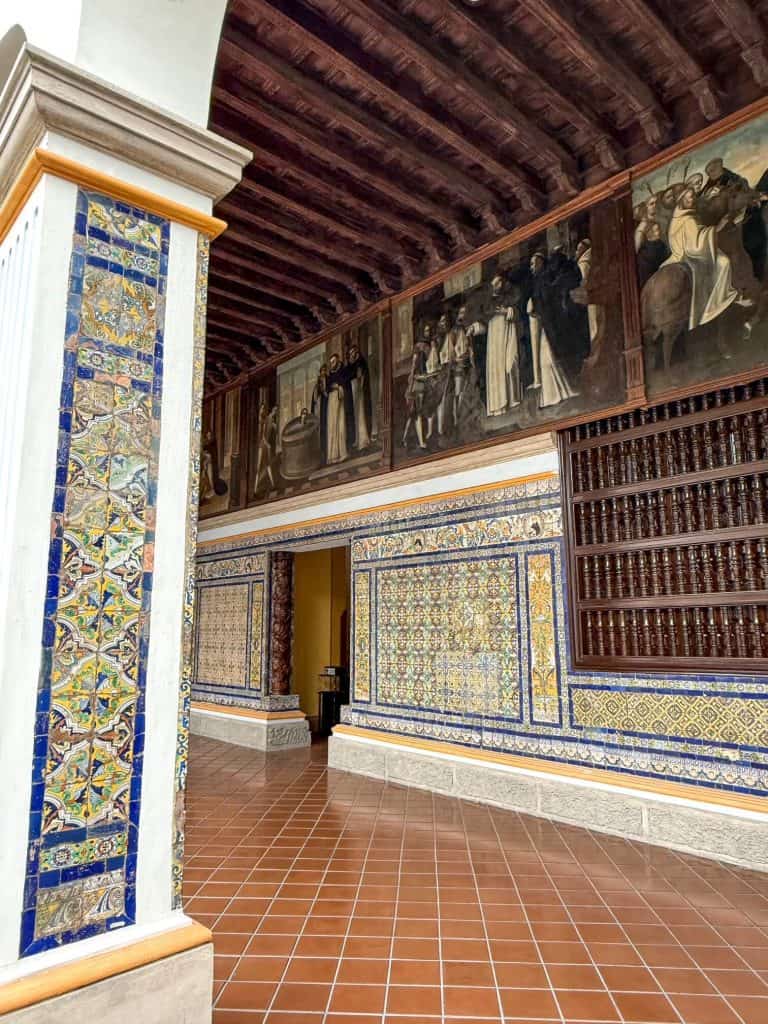 View of the intricate tiles in blue, yellow and white along the walls and pillars of the cloisters bordering the courtyard with a series of painting on the top panels above the tiles at Santa Domingo Convent in Lima.