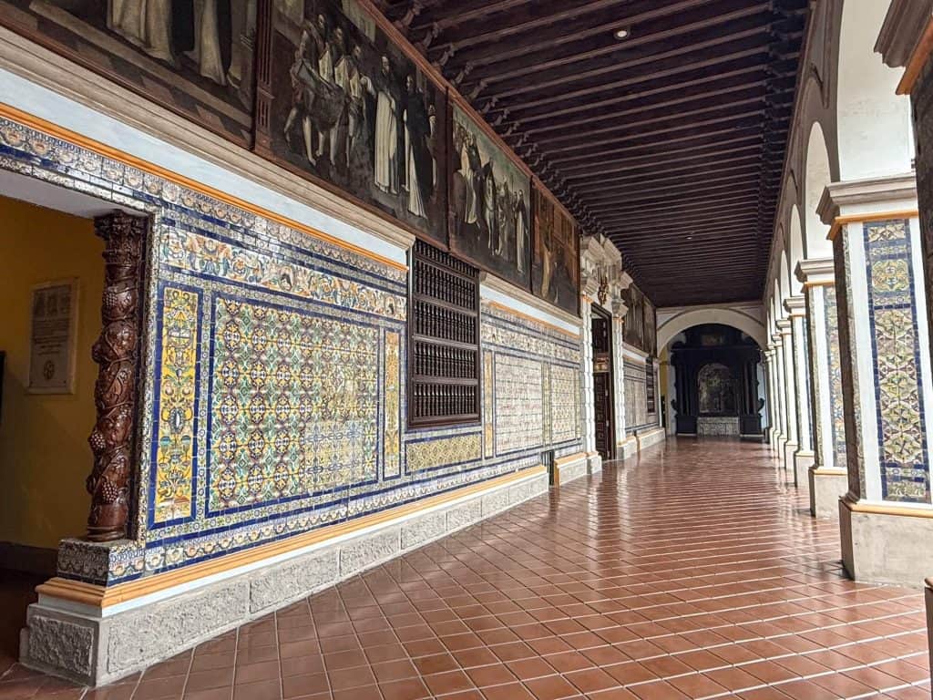 Looking down the hall of the cloisters with yellow, blue and white tiles on the lower half of the walls and paintings on the top portion in Spanish-Moorish styles at Santa Domingo Convent.