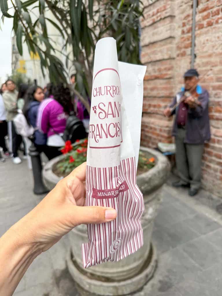 Holding up the white and red bag with a churro inside at Churro San Francisco with a man playing an instrument in the background against a brick wall.