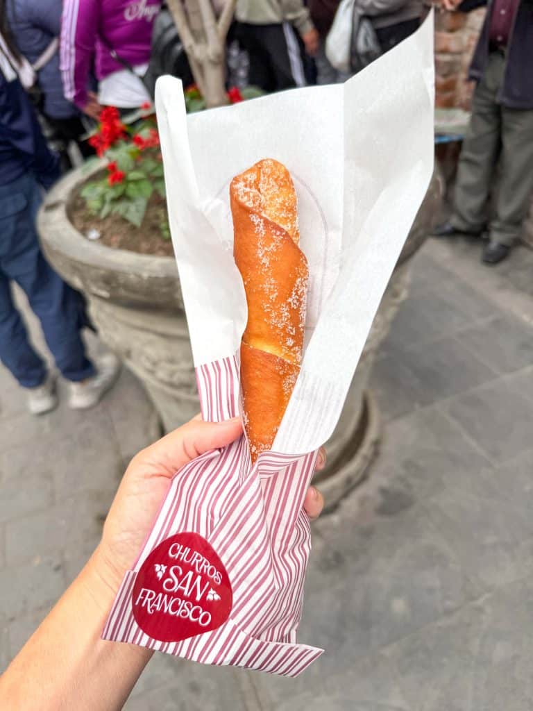 A large churro filled with Manjar blanco filling in a white and red bag with a sticker on it that says Churros San Francisco near the San Francisco Church in Lima.