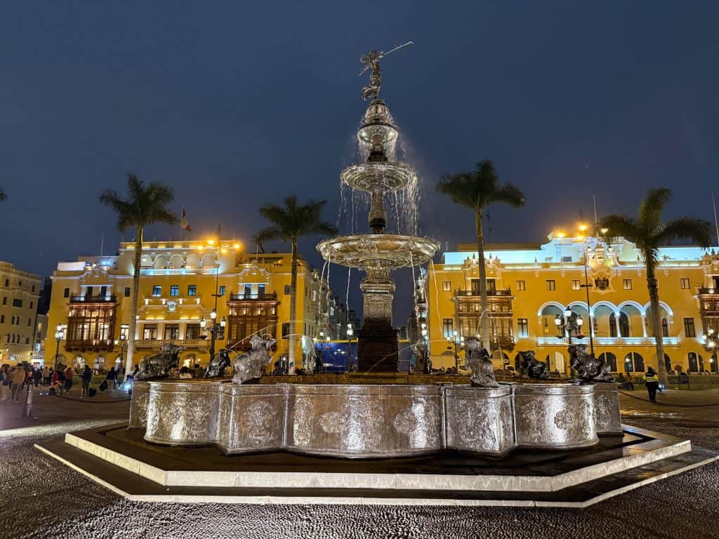 A beautiful water fountain in the center of Plaza Mayor in Lima at night with the lighting illuminating the craftsmanship around the fountain palm trees and yellow buildings in the backdrop.