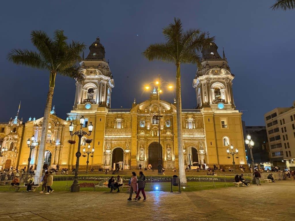 The Lima Cathedral lit up at night with Plaza Mayor in front lined with palm trees and people walking by.