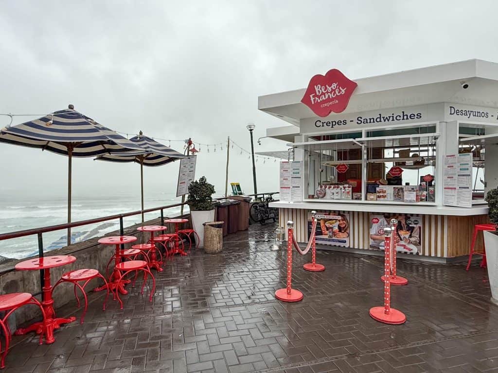 View of Besos Frances, a cute white and red stand along the cliffs overlooking the Pacific Ocean with red chairs and blue and white striped umbrellas that serves crepes in Lima.