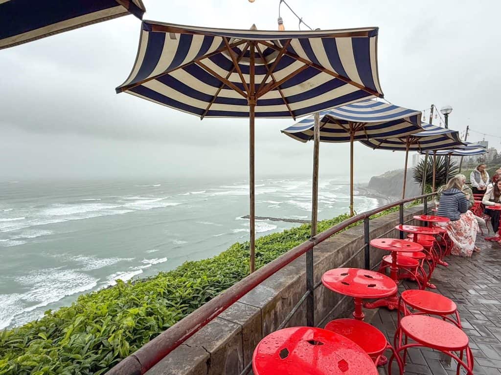 Looking out over the ocean with a row of red stools, blue and white striped umbrellas with people sitting and eating crepes on a foggy and overcast day in Lima.