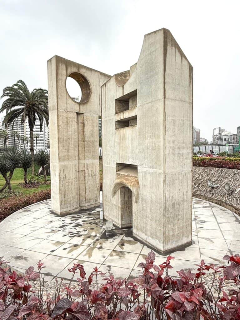 A concrete statue with abstract shapes that is around 15 feet tall on a circular base at the start of the pathway of Malecon Miralfores in Lima along the coast.