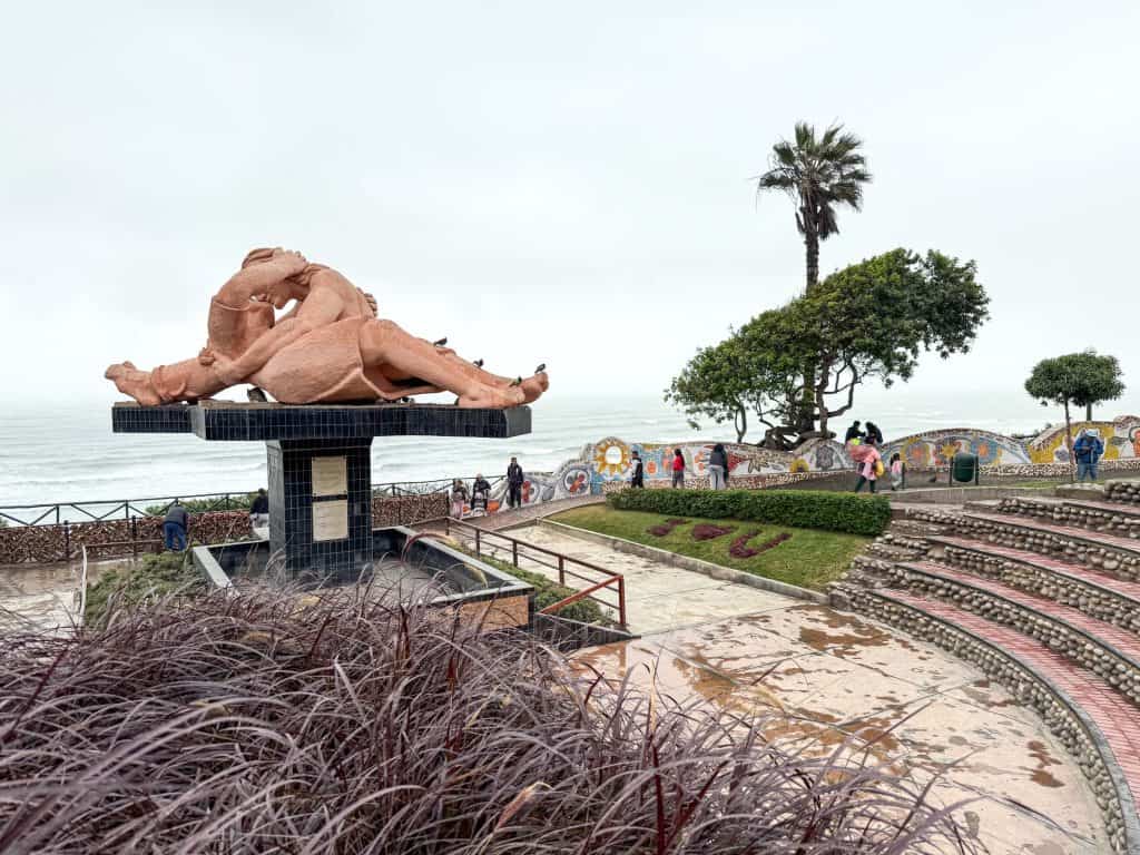Standing almost behind the Lover's Kiss statue at Love Park with a small amphitheatre seating with palm trees and a direct view of the Pacific Ocean on a cloudy day in Lima.