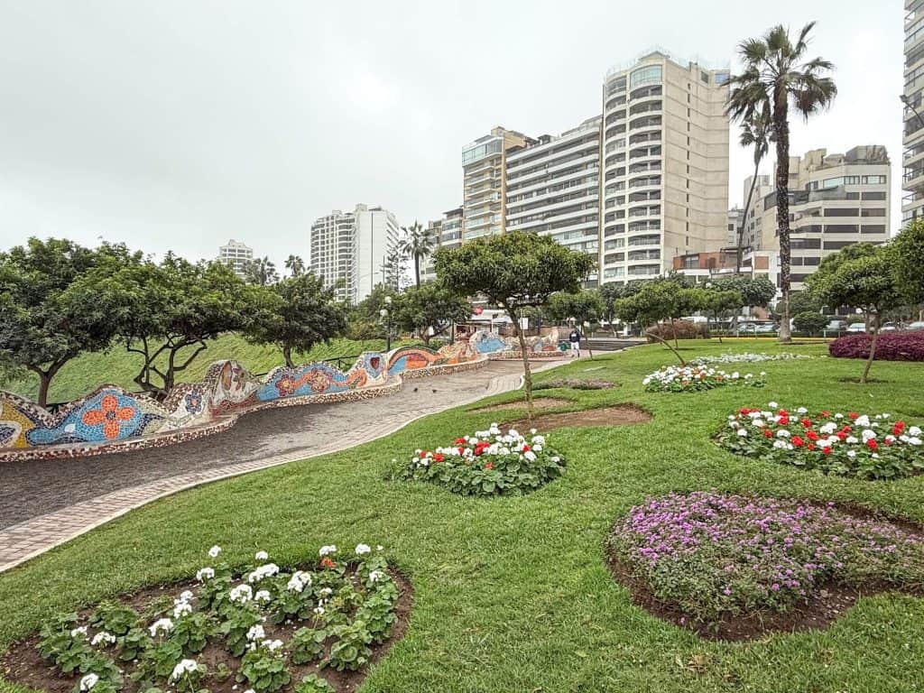 A wide view of the grassy park with white and purple flowers, trees, and curving wall made up of tiles creating designs and quotes at Love Park in Lima.