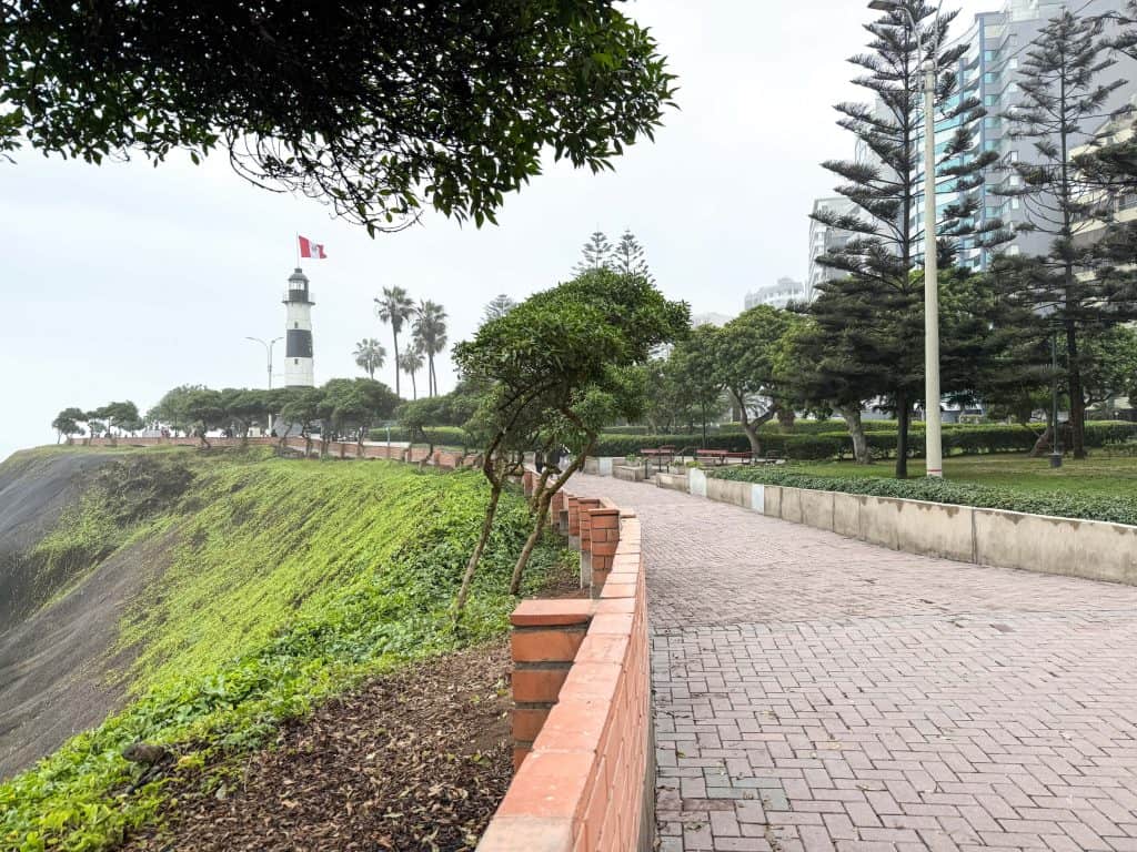Walking on the paved pathway of Miraflores Malecon bordered by trees and incredible view of the ocean with a lighthouse at end of the path.